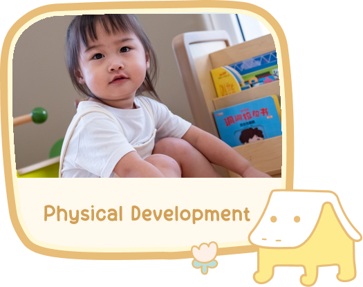 Physical Development — a toddler sitting on the floor beside a book rack at PikoPoko, knees drawn up.
