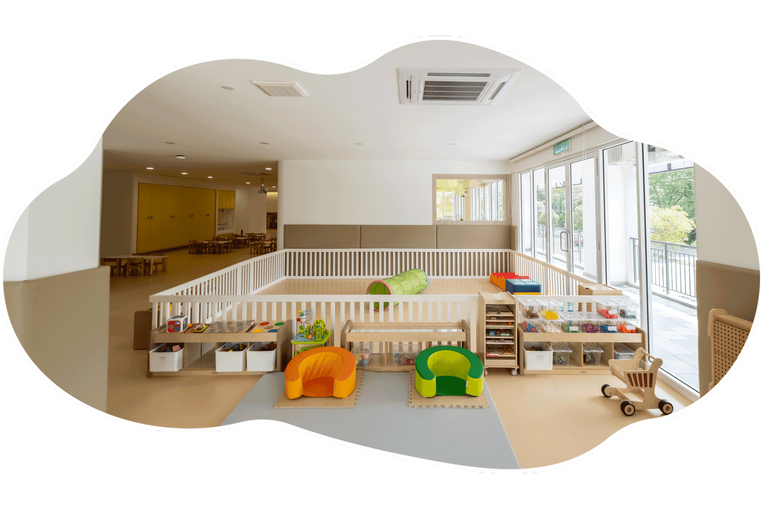 A play area at the centre, with a low-fenced soft-play zone, open shelves of toys and a wall of windows.